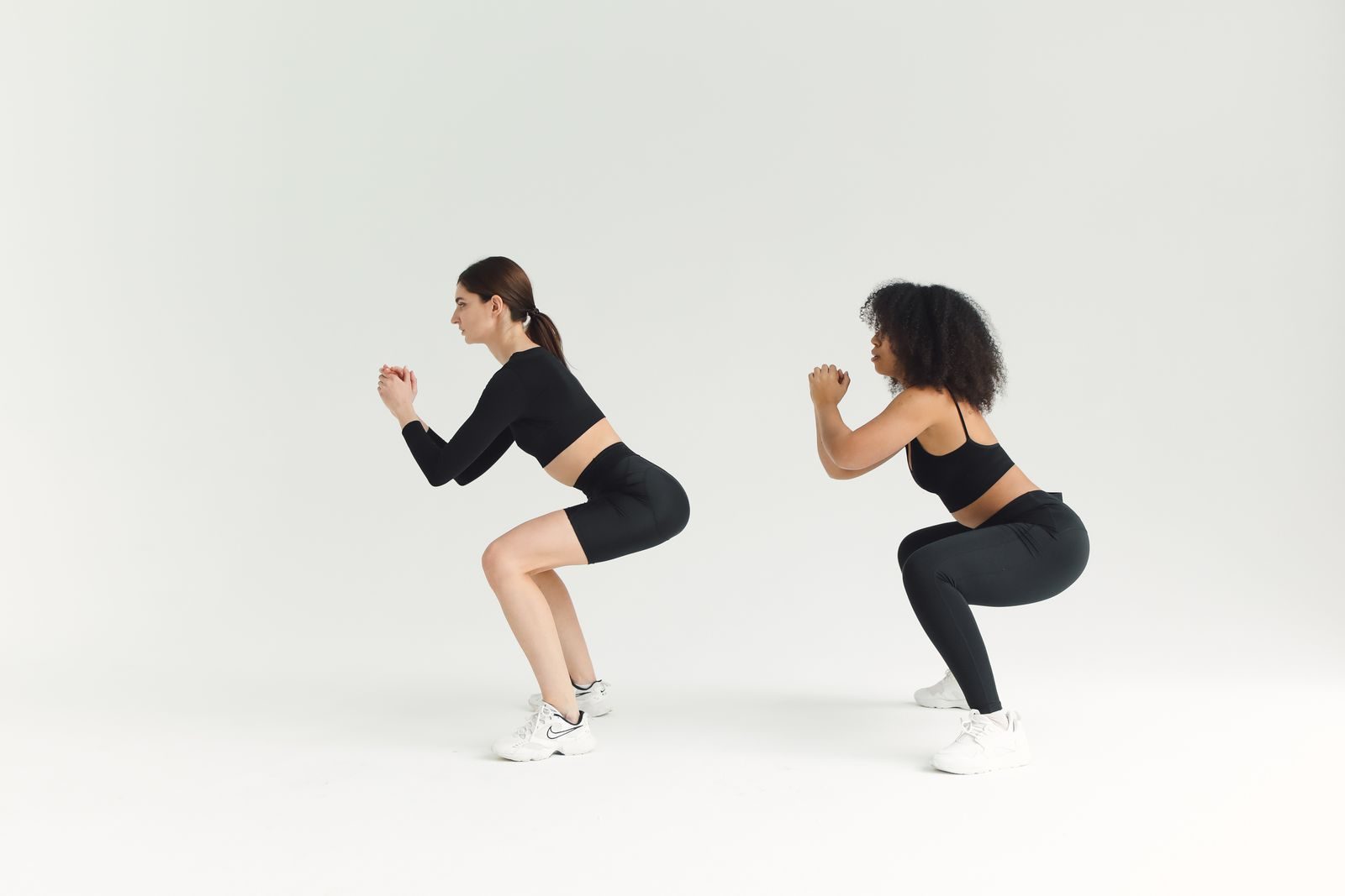 Two women performing squats in sportswear against a plain background, emphasizing fitness and a healthy lifestyle.