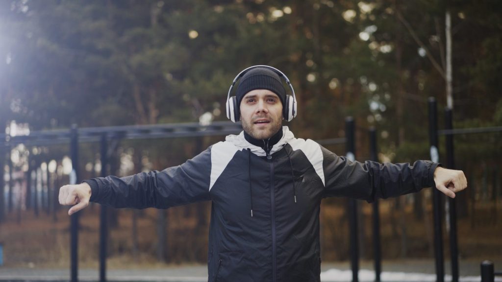 Man with headphones stretching outdoors with arms outstretched.
