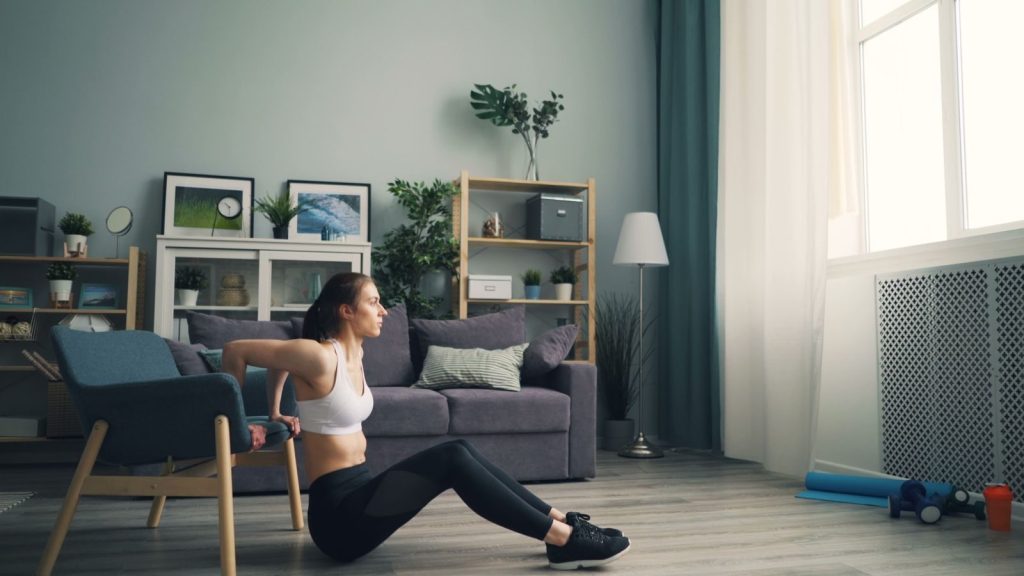 Woman doing tricep dips at home using a chair for support, promoting fitness indoors.