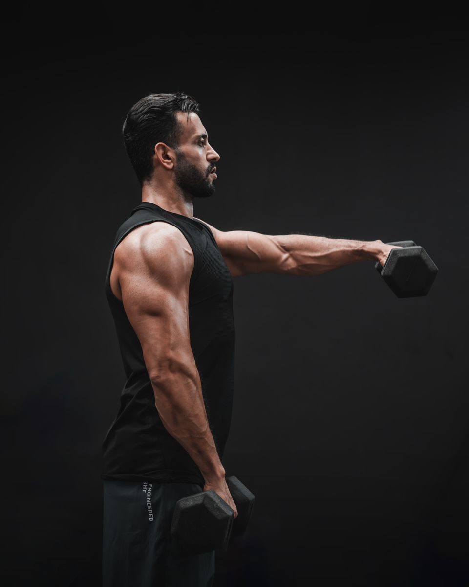 Side profile of a muscular man working out with dumbbells in a gym setting.