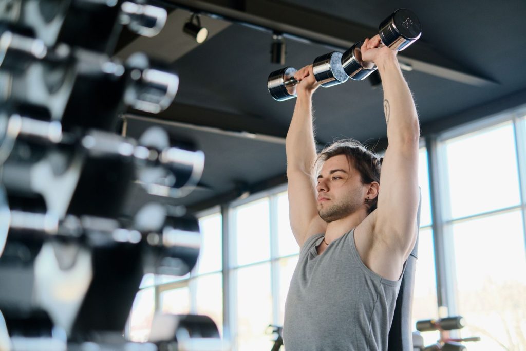 Man performing shoulder press with dumbbells in a well-lit gym environment.