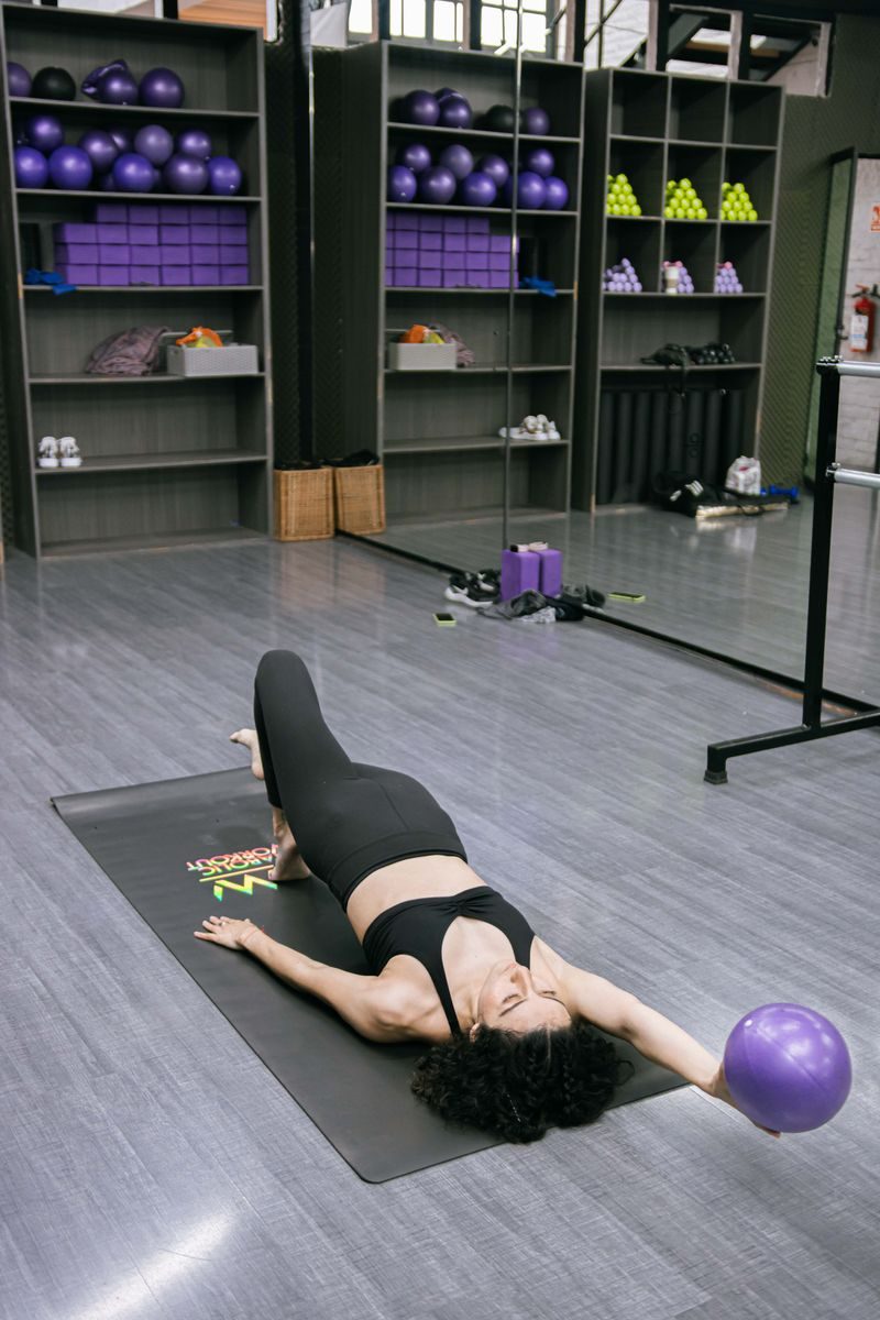 A woman in sportswear exercising with a ball on a mat in a gym setting.