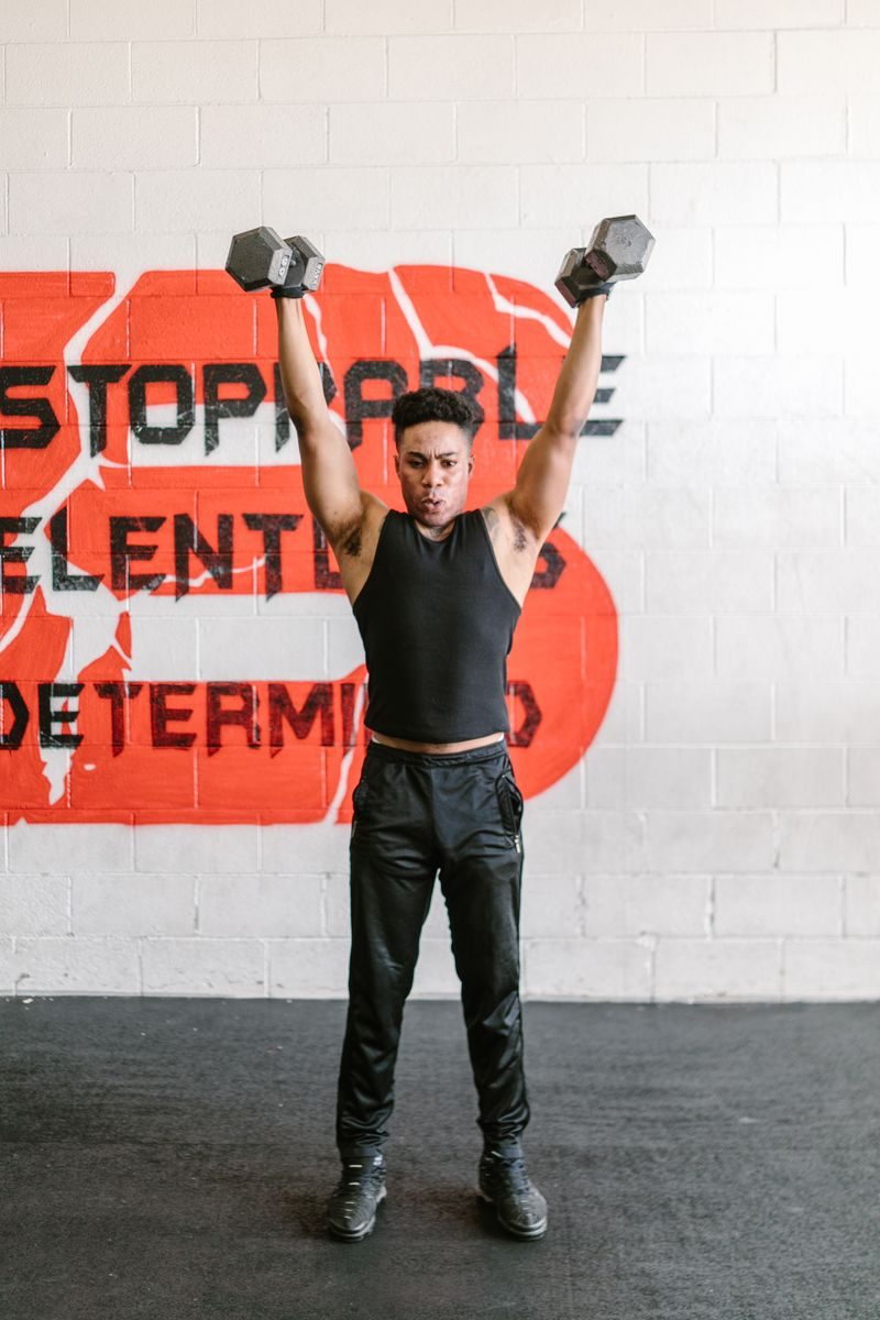 African American man lifting dumbbells overhead in a gym with motivational wall art.