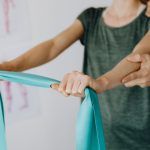 Crop unrecognizable orthopedist in uniform and wristwatch supporting shoulder and reached arm of faceless female patient with resistance band in doctor office
